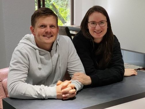 Dimi und Rebecca sitzen lächelnd nebeneinander am Tisch