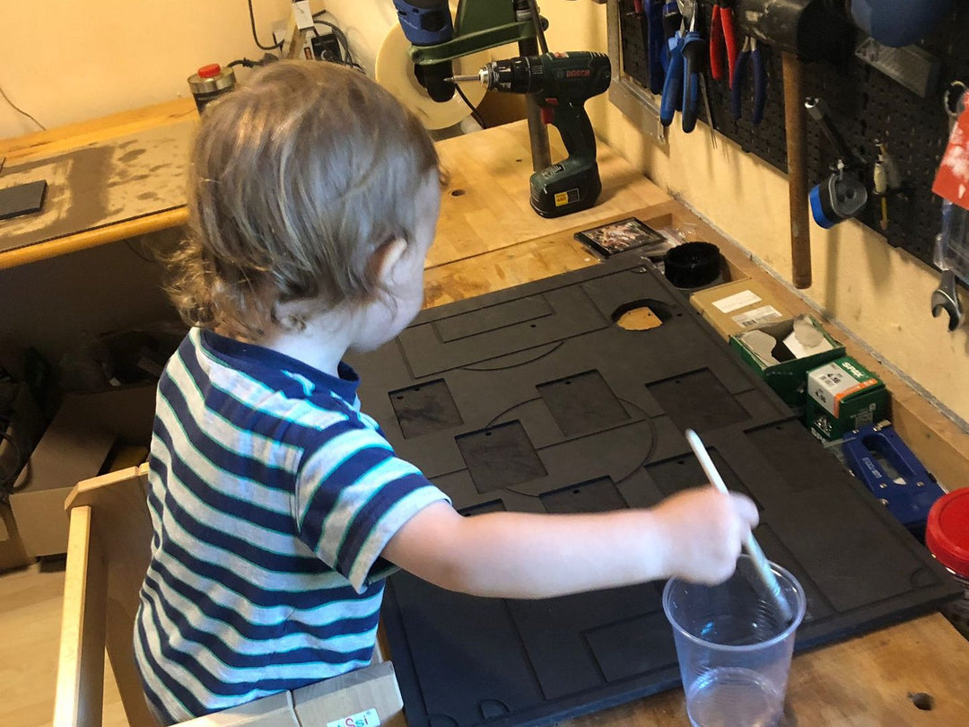 Kleiner Junge in gestreiftem Shirt pinselt in der Werkstatt mit Wasser über einen Formationsrahmen